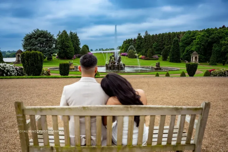 A couple sitting on a wooden bench, enjoying a serene view of a beautifully landscaped garden with a grand fountain in the distance. Photo by Royal Bindi Photography.