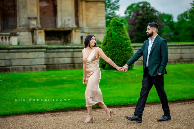 Couple candid moment holding hands passing a historic building in a green garden.