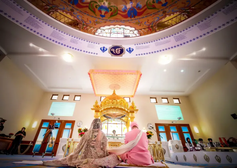 A bride and groom, dressed in traditional Indian wedding attire, sit together in a gurdwara (Sikh temple) during their wedding ceremony. The couple is positioned in front of a richly decorated altar with a golden canopy, under which a Sikh priest (Granthi) is seated. The bride wears a beautifully embroidered lehenga, while the groom is dressed in a pink sherwani and turban. The interior of the gurdwara is adorned with floral arrangements and traditional decorations, with cameras set up for capturing the event.