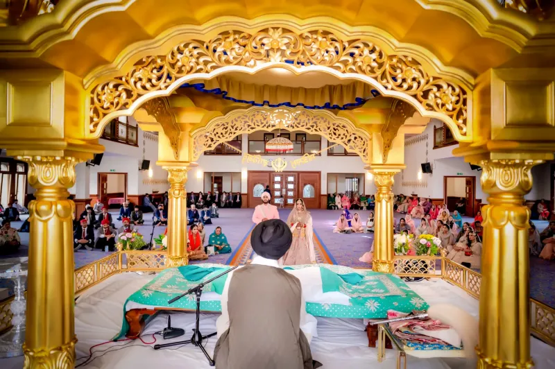 Sikh Wedding Ceremony in Gurdwara: Bride and Groom Stand Before the Granthi during Anand Karaj. The Gilded Decor of the Gurdwara\'s Interior Enhances the Setting, Surrounded by Seated Guests in Traditional Attire. 