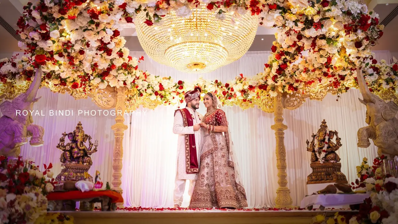 A beautifully decorated wedding ceremony with the couple standing in front of a traditional Indian mandap. The mandap is decorated with flowers and gold accents and there are two statues of the Hindu deity Ganesha on either side of the couple. The image is captured by Royal Bindi Photography.