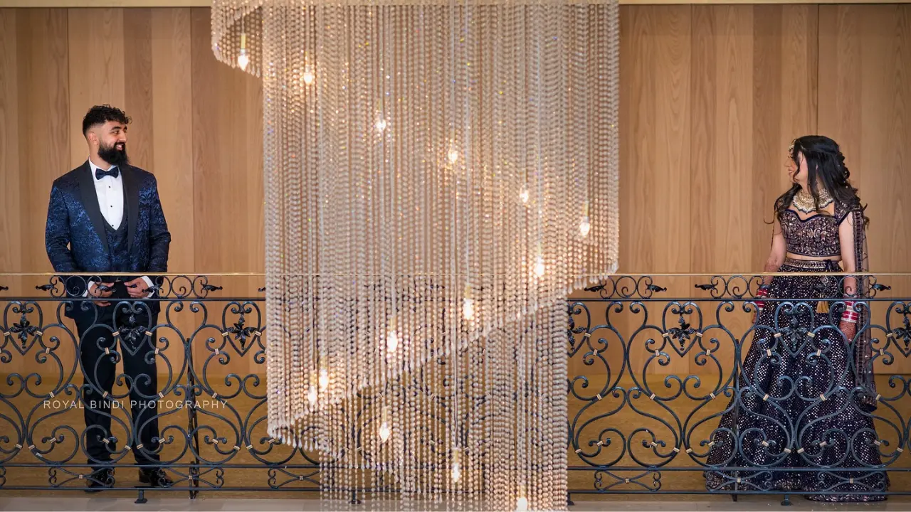 A bride and groom stand on opposite sides of a balcony, separated by a sparkling crystal chandelier. The bride is wearing a beautiful navy blue and gold lehenga, and the groom is in a navy blue suit with a black bow tie. The couple looks at each other with love and joy.