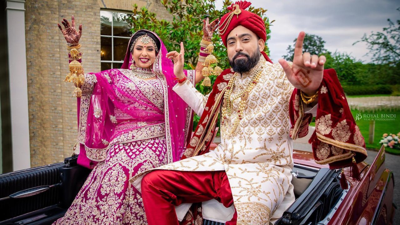 A newly married couple, the bride in a vibrant pink and gold lehenga and the groom in a traditional white kurta, are beaming with joy and excitement as they ride in a car. They are both making peace signs with their hands, showing their love for each other and their excitement for their new life together. The image is a vibrant celebration of Indian culture and tradition, capturing the joy and happiness of a wedding day.