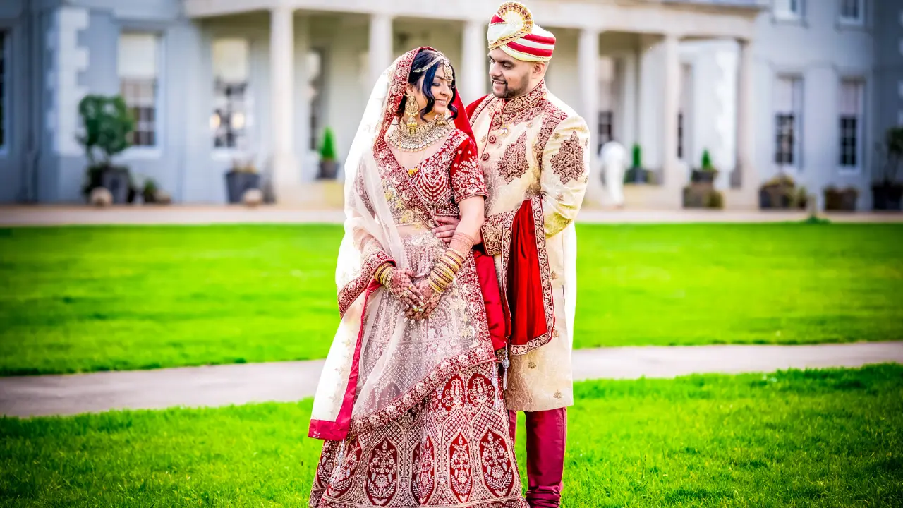 A bride and groom in traditional Indian wedding attire stand on a grassy lawn. The groom is wearing a beige sherwani with a red turban, while the bride is wearing a red and gold lehenga with a white veil. They are looking at each other lovingly.