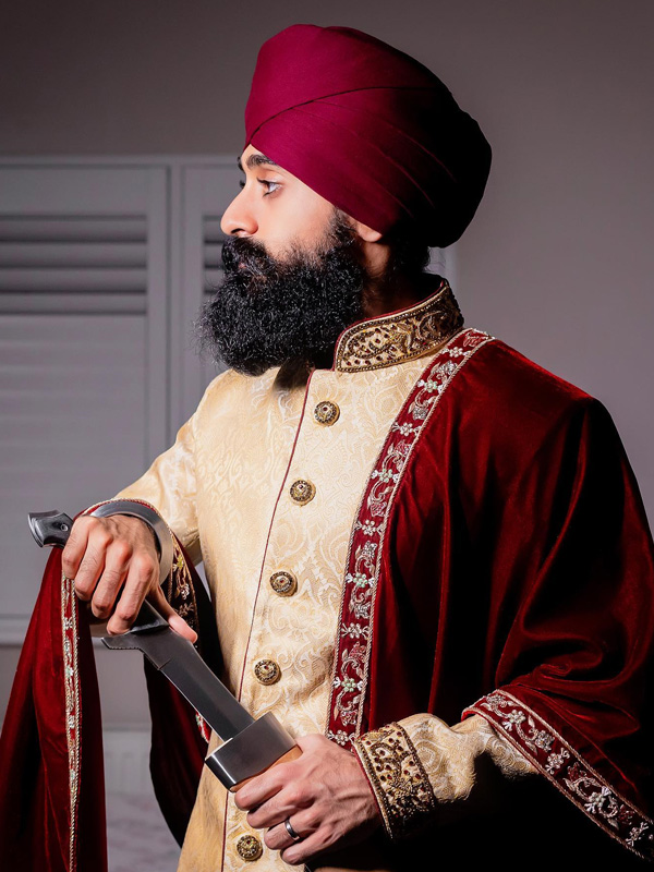 Sikh man in a cream and maroon achkan holding a sword.