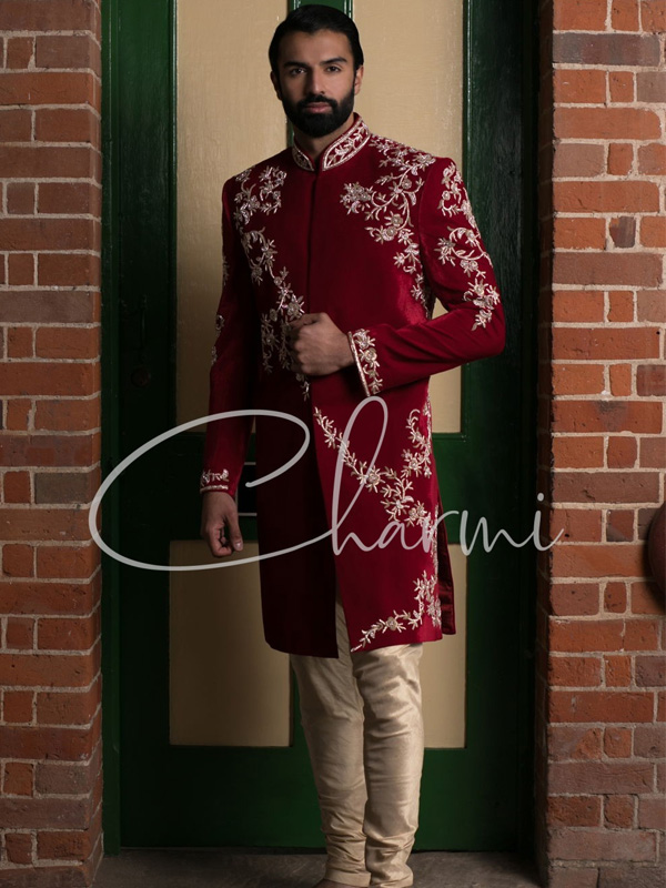 A man wearing a red embroidered sherwani with golden floral patterns, standing in front of a green and beige door.