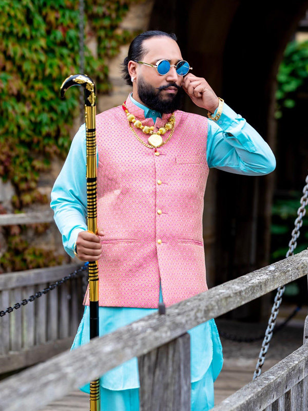 Man in a pink waistcoat and blue kurta with sunglasses and a cane.