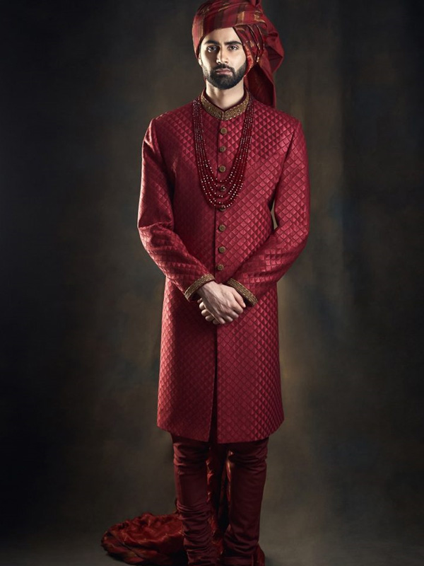 A man wearing a red, intricately patterned sherwani with a matching turban and red beaded necklaces, posing elegantly in traditional Indian wedding attire.