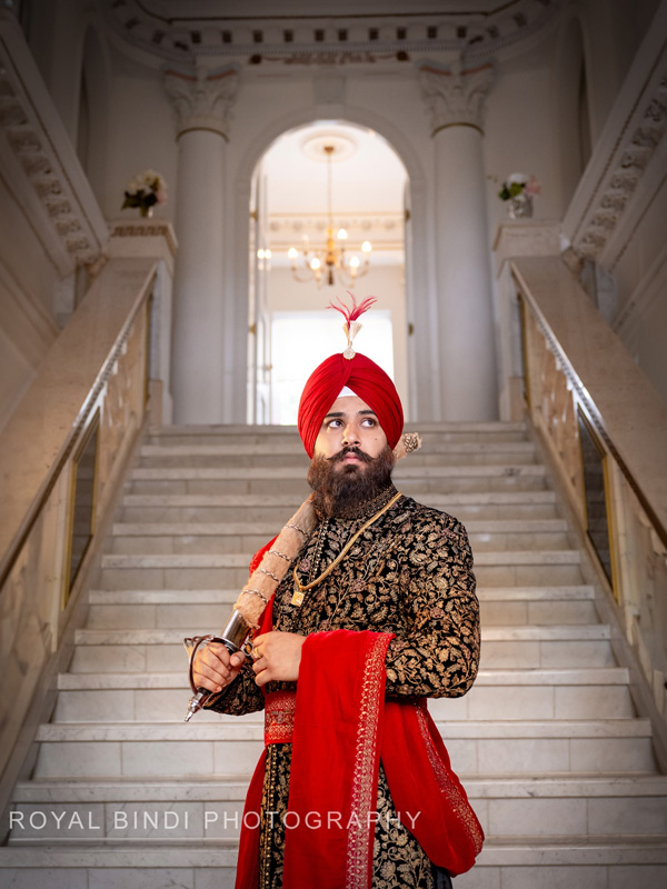 Man in a red turban and embroidered sherwani holding a sword on stairs.