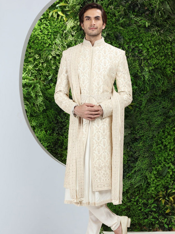 A man in an elegant cream sherwani stands in front of a lush green backdrop.