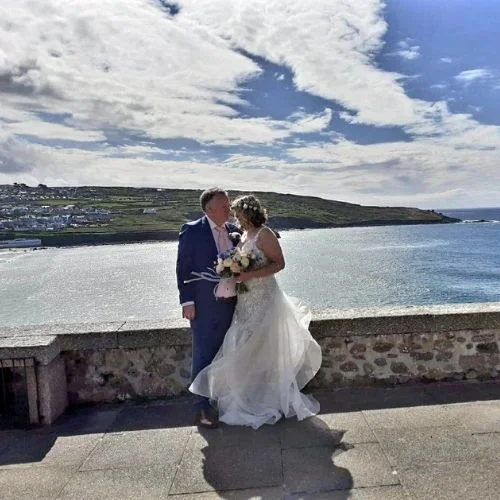 A bride and groom standing by the ocean, sharing a moment with the sea and coastal landscape in the background.