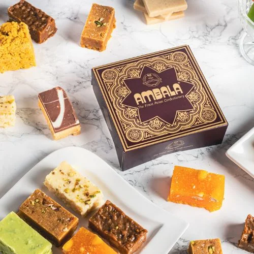 A square, maroon box with gold designs and the brand name "Ambala" on it, containing various Indian sweets arranged on a marble surface.