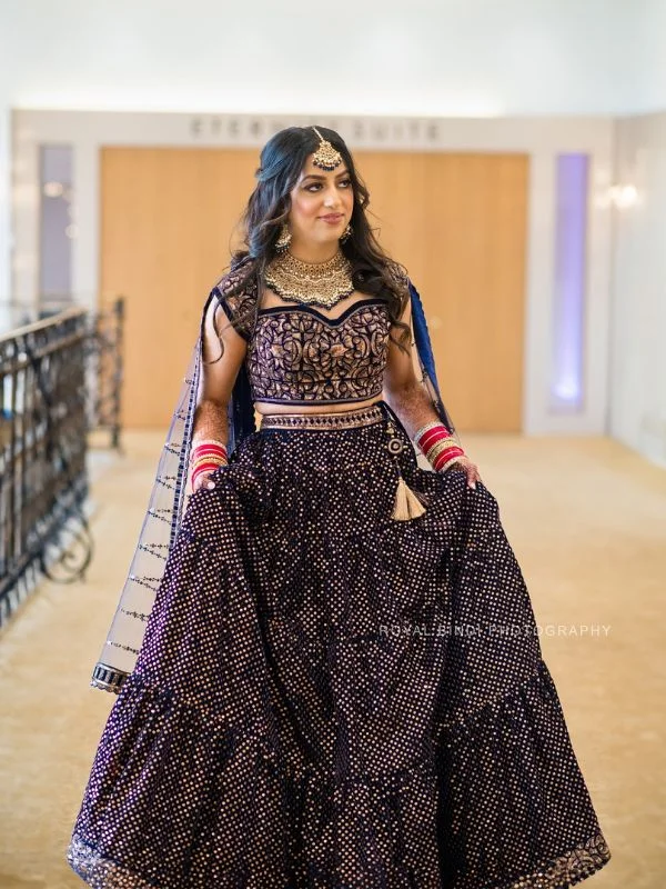 Woman in navy lehenga with intricate designs, wearing a belted dupatta and traditional jewellery.