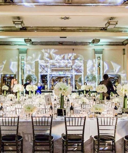 A photo of a grand ballroom set up for a formal event. Long tables are adorned with white tablecloths, centerpieces, and glassware.