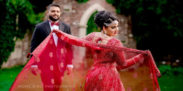 A bride wearing an elegant red embellished dress and jewelry, playfully holding her dupatta, with a groom in a black suit and red bow tie smiling in the background.