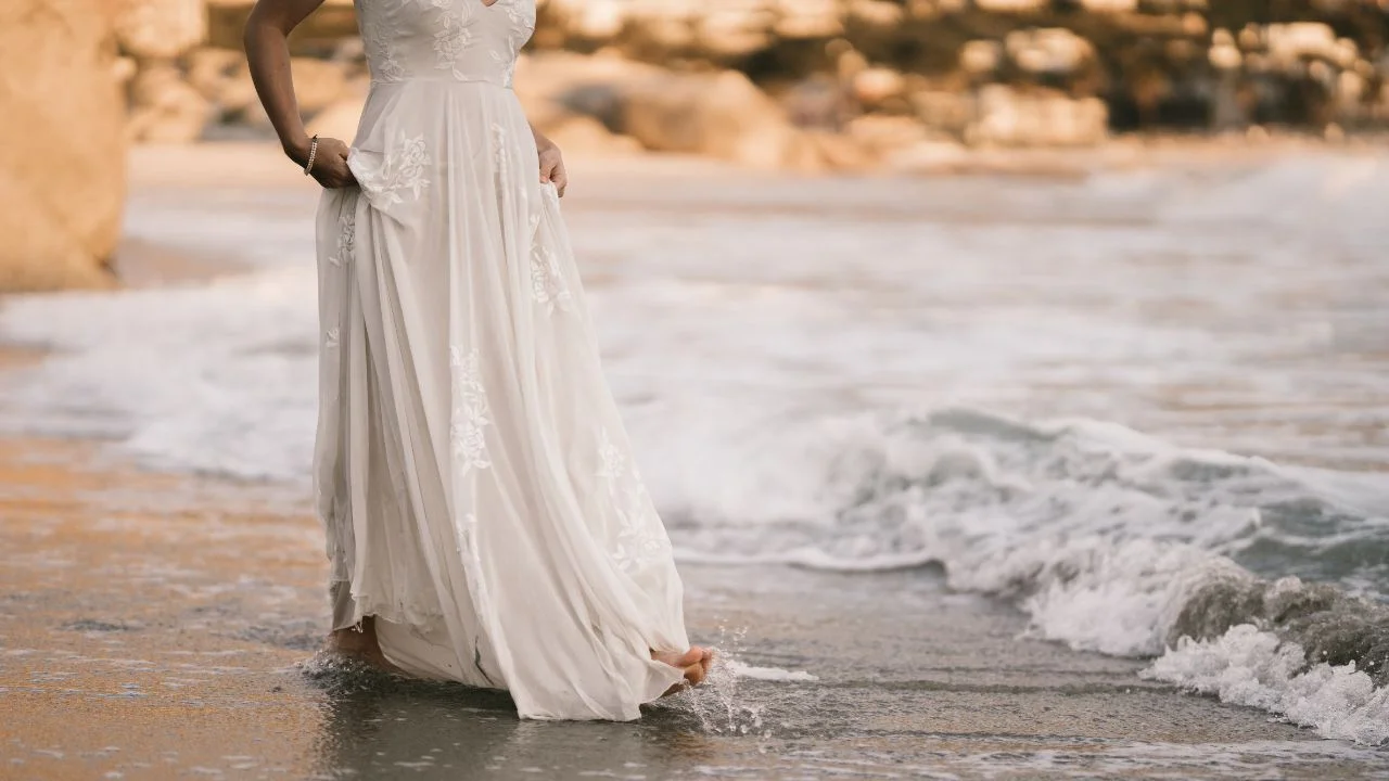 A bride wearing a flowing white wedding dress walking barefoot along the beach with gentle waves touching her feet.