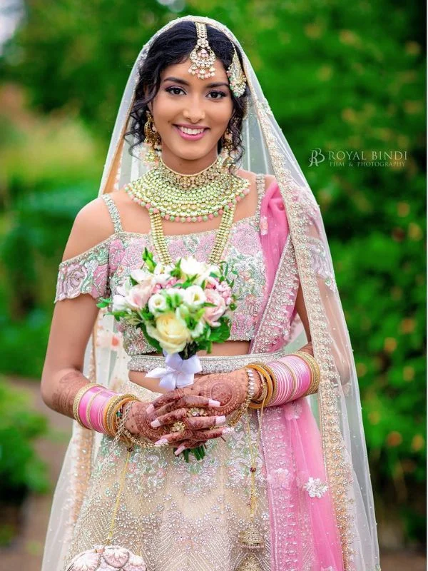 Bride in pink and ivory lehenga with cape-style dupatta, holding a bouquet and wearing traditional jewelry.