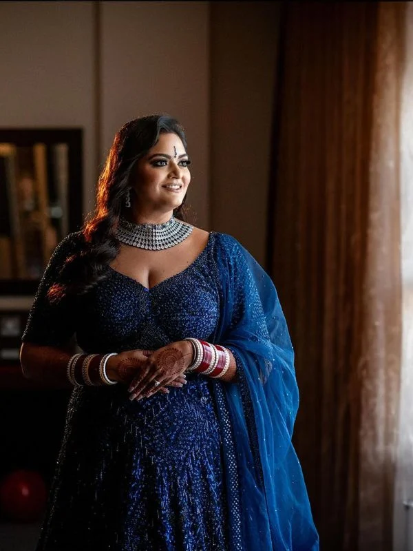 Bride in glittery blue dress and diamond necklace, standing by window.