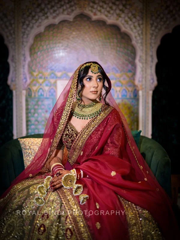 Bride in red lehenga with embroidered dupatta and heavy traditional jewelry, seated in ornate setting