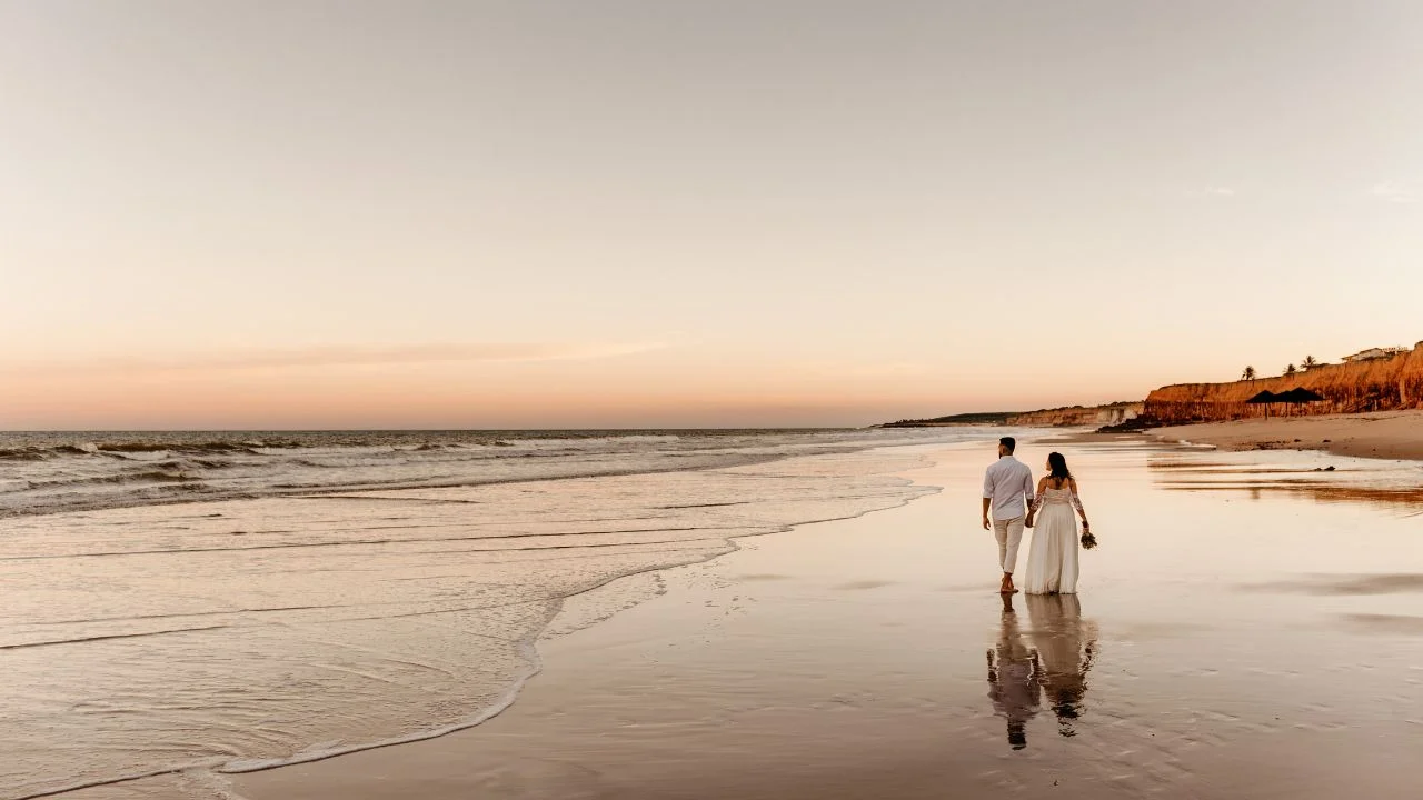 A bride and groom walking hand in hand along a peaceful beach at sunset, with the golden sky reflecting on the wet sand and gentle waves in the background.