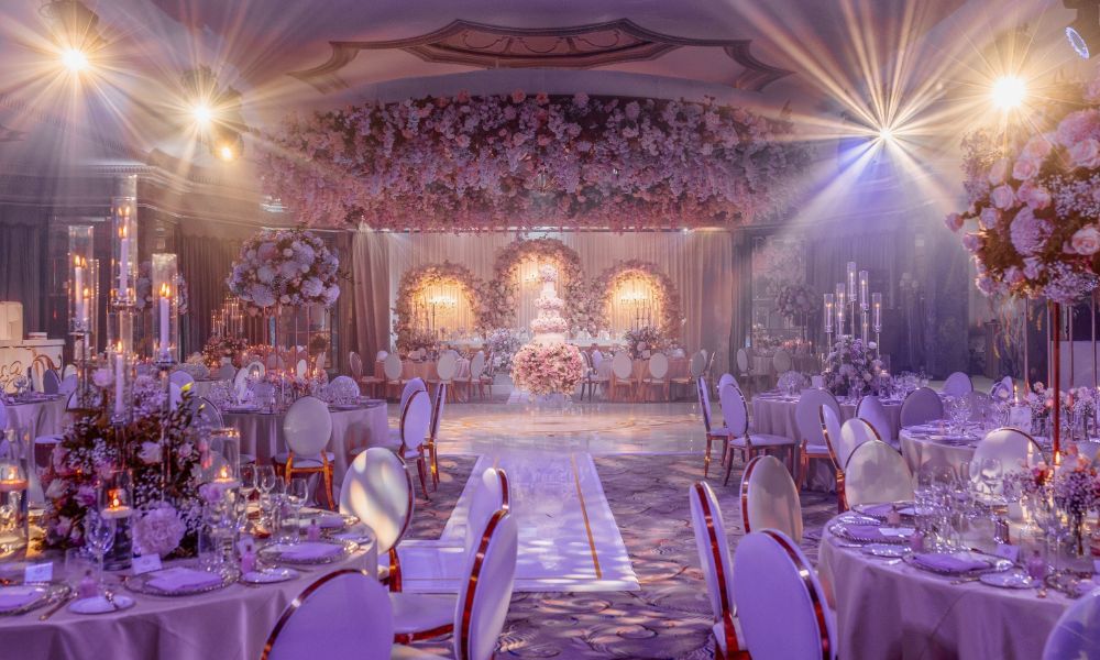 A grand ballroom at The Dorchester Hotel in London, decorated for a lavish wedding reception. The room features a stunning floral backdrop, a tiered wedding cake, round tables with white tablecloths and floral centerpieces, and a white runner leading to the dance floor.