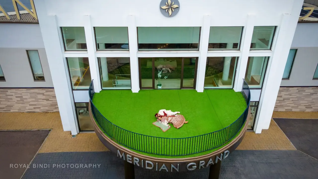 Indian bride and groom in traditional wedding attire lying on the green balcony of Meridian Grand, photographed from a top view by Royal Bindi Photography.