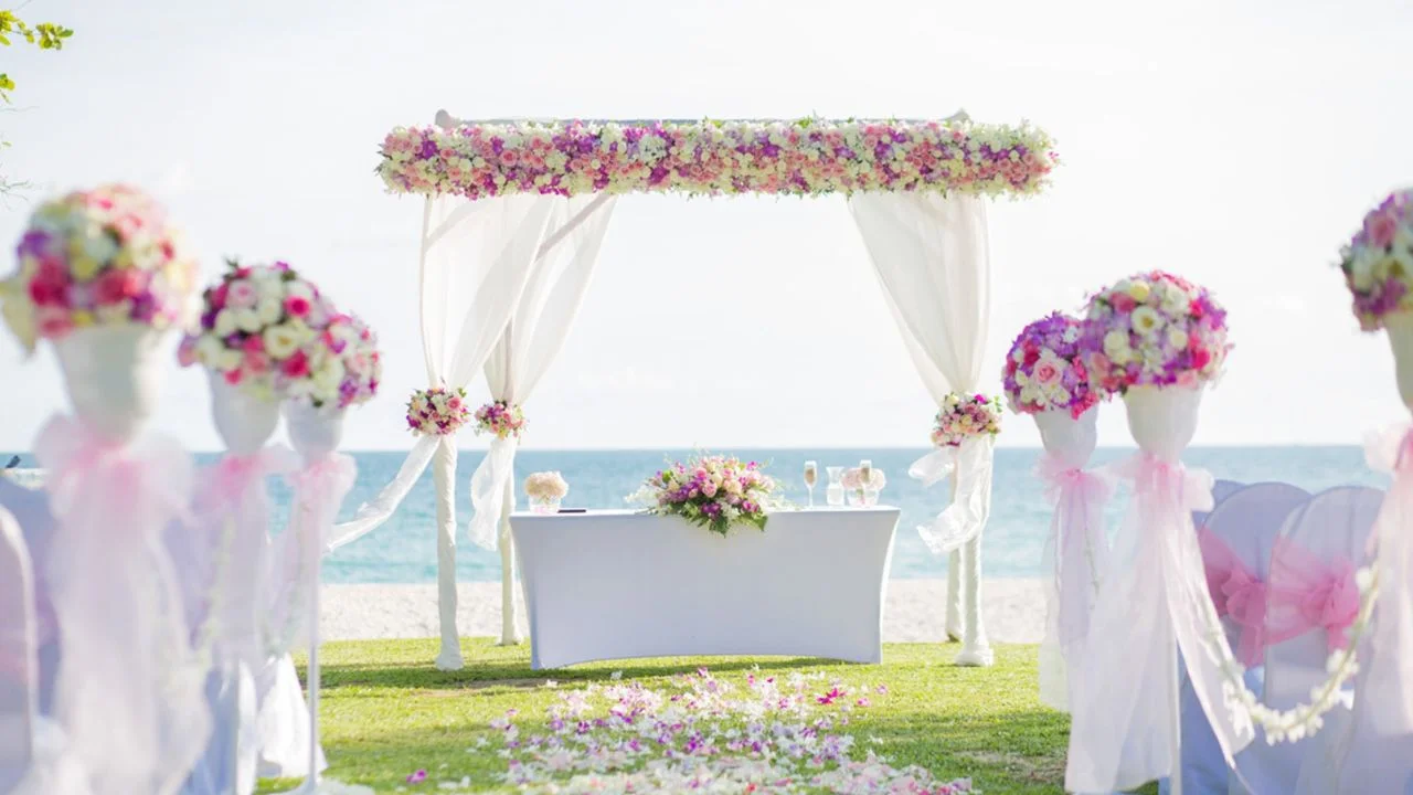 A beach wedding setup with a floral arch, table, and chairs.