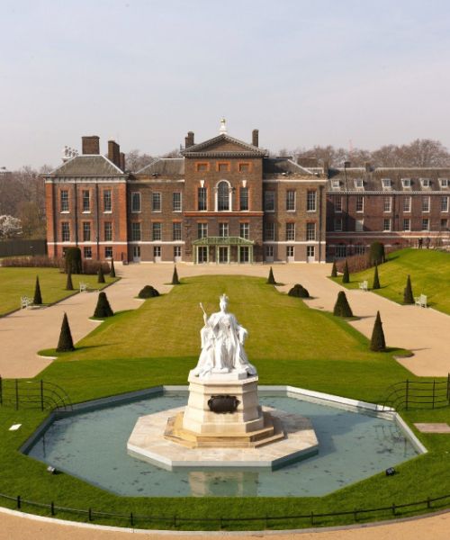 Kensington Palace, a royal residence in London, England. The palace is surrounded by green lawns and gardens, with a statue in the foreground.