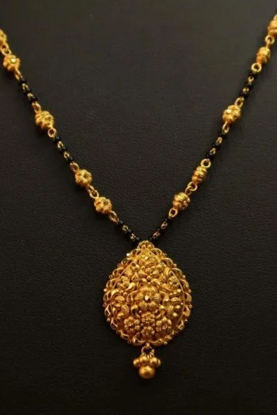 A close-up image of a traditional gold mangalsutra with a round pendant adorned with intricate floral designs. The pendant is suspended from a chain made of black beads.