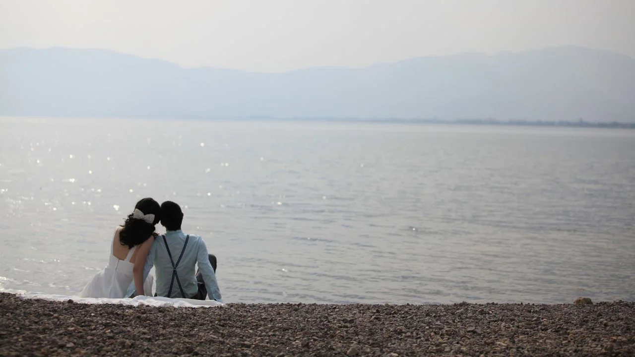 Back view of a bride and groom sitting together by a peaceful lakeshore, embracing a romantic moment.