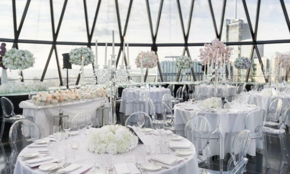 Wedding reception setup with round tables, white tablecloths, and floral centerpieces. Large windows provide breathtaking views of the city skyline.