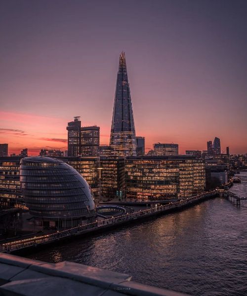 A panoramic view of London's skyline at dusk. The iconic Shard dominates the scene, illuminated against a vibrant pink and purple sky. The River Thames and other city landmarks are visible in the background.