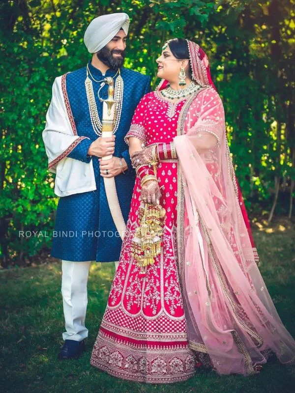 Sikh couple smiling, bride in red lehenga and groom in blue sherwani outdoors.