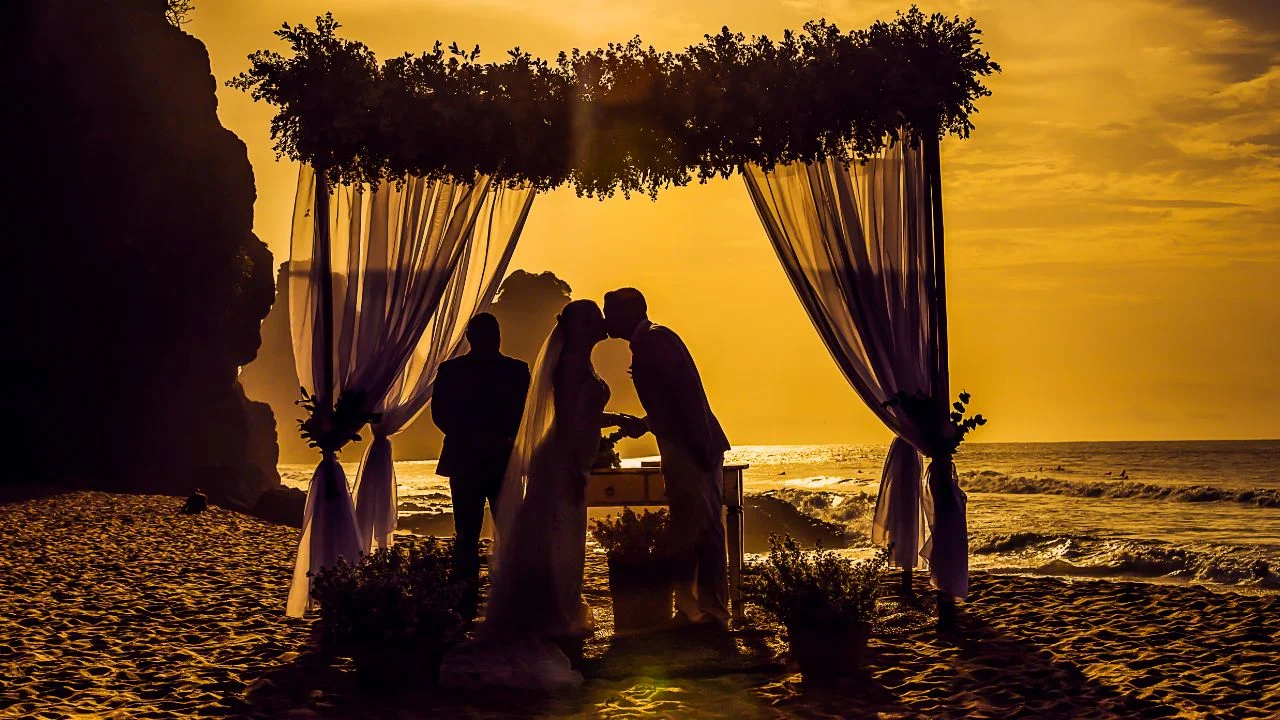 Bride and groom kissing under a floral archway during their beach wedding ceremony at sunset, with the ocean in the background and silhouettes of the officiant and couple against the golden sky.