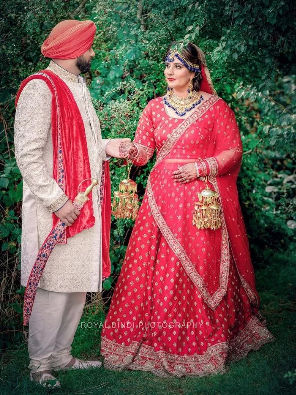 Sikh couple in red lehenga and ivory sherwani holding hands outdoors.