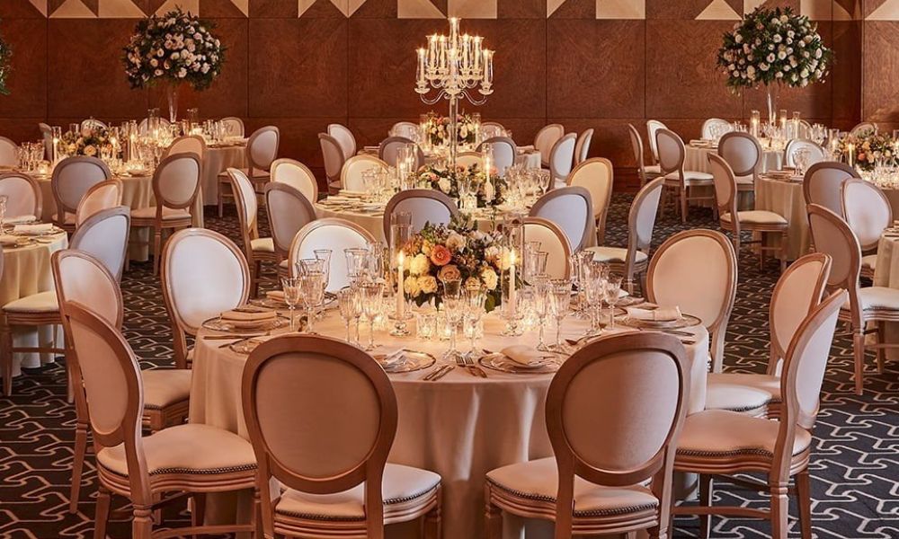 A photo of a grand ballroom set up for a formal event. Round tables are adorned with white tablecloths, centerpieces, and glassware.