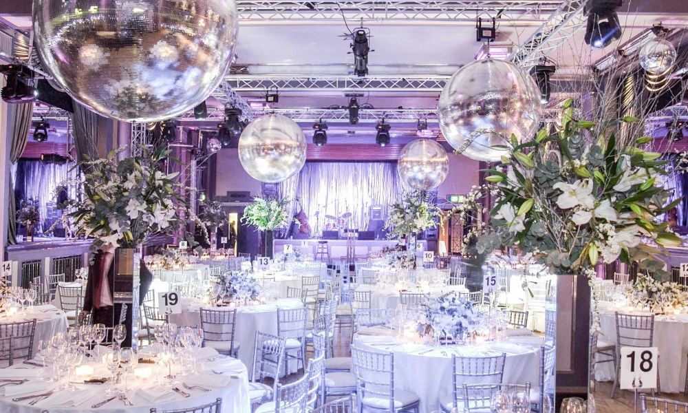 A photo of a grand ballroom decorated for a festive event. Round tables are adorned with white tablecloths and centerpieces, and a large disco ball hangs from the ceiling.