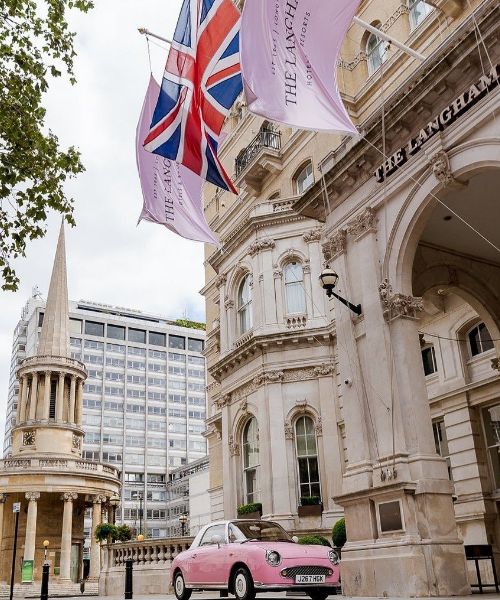 A photo of The Langham hotel in London, England. The hotel is a grand building with a pink car parked in front and Union Jack flags flying.