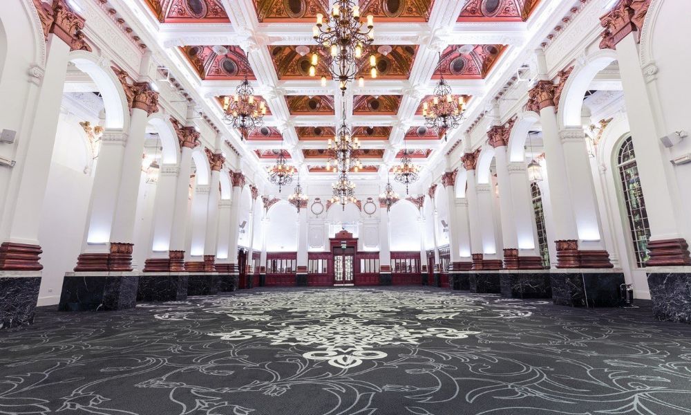 A photo of a grand ballroom with high ceilings, ornate columns, and a patterned carpet.