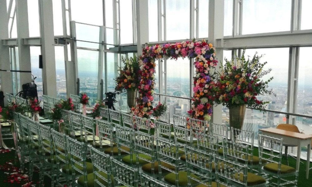 A wedding ceremony setup at The Shard in London, England. The venue features floor-to-ceiling windows with panoramic city views, a floral arch, and clear chairs arranged for guests.