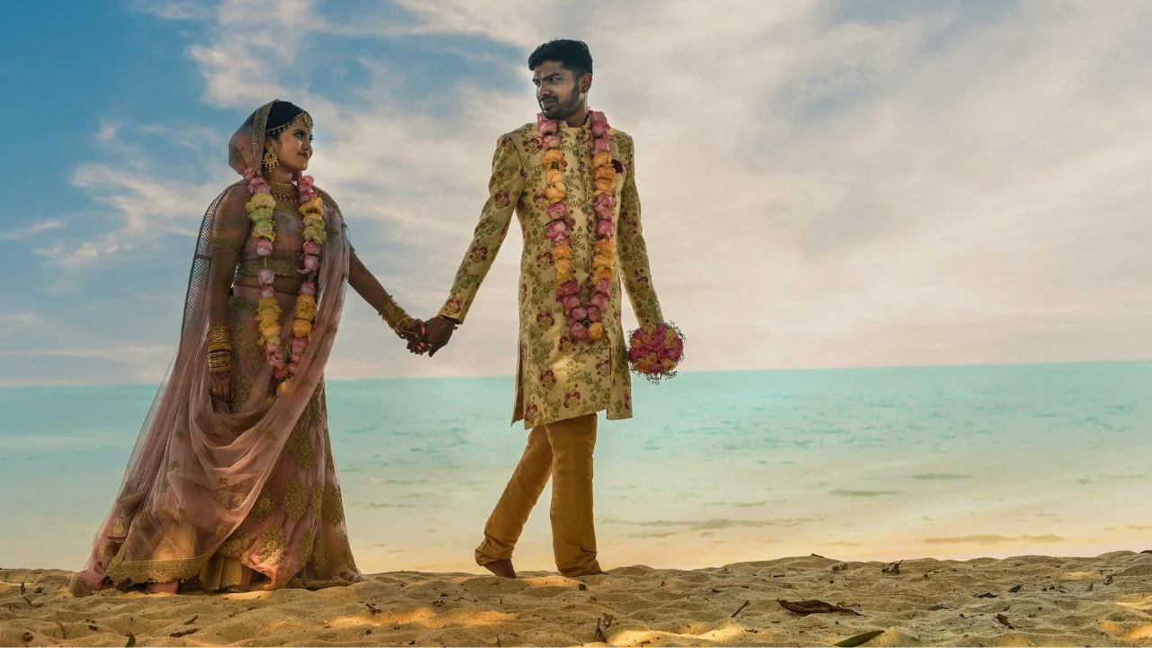 Bride and groom in traditional Indian wedding attire, adorned with floral garlands, holding hands and walking on a beach under a bright sky.