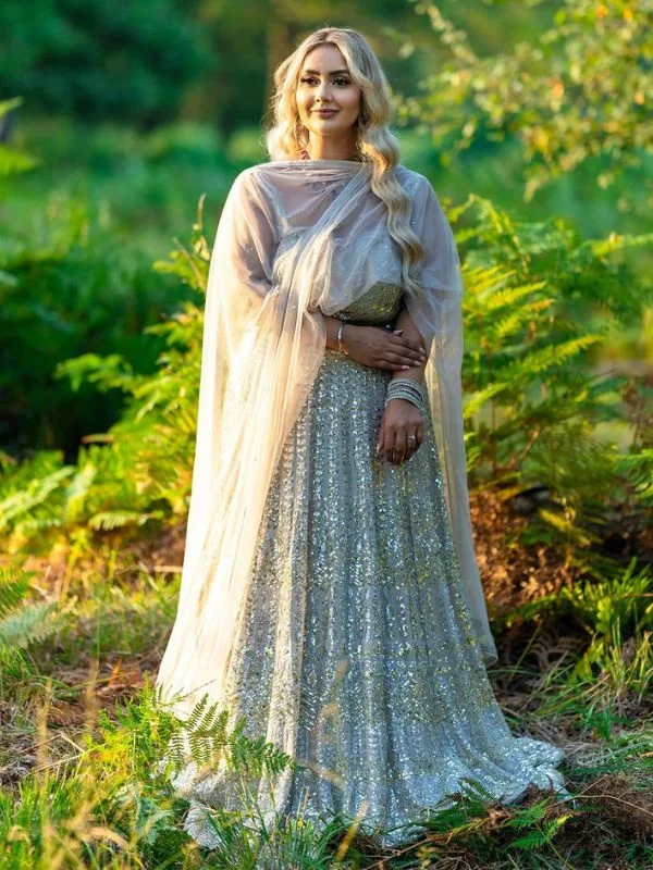 Woman wearing a sparkling silver gown with a sheer cape, standing in a green forest setting.