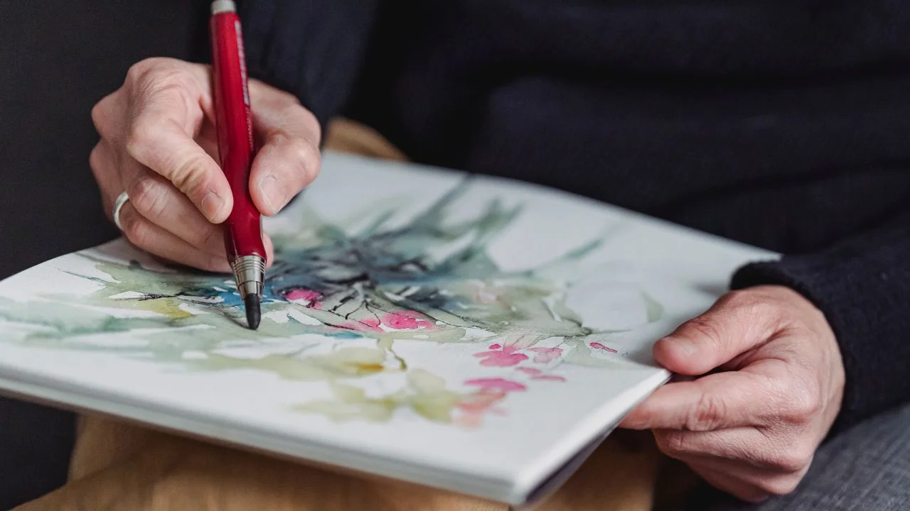 A close-up of a person's hands holding a paintbrush and working on a watercolor painting with abstract floral details on paper.