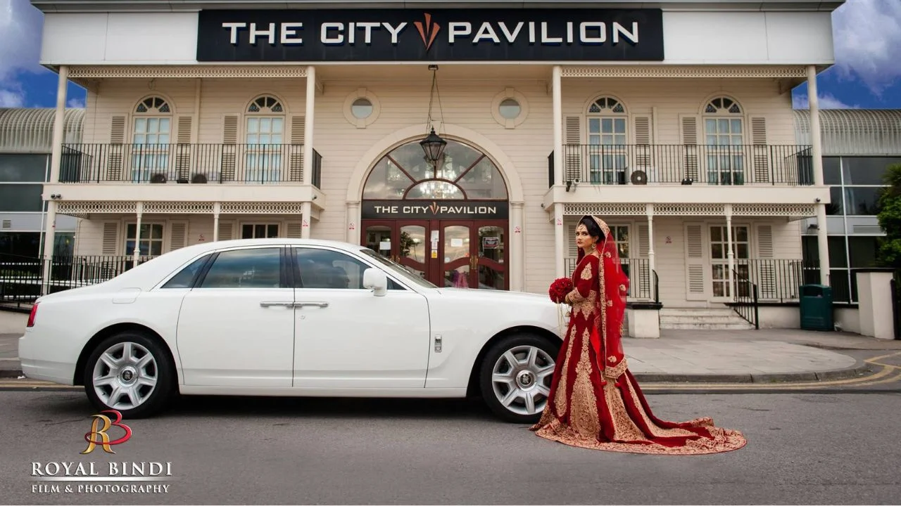 Asian bride in red traditional wedding attire posing next to a white luxury car in front of The City Pavilion venue.