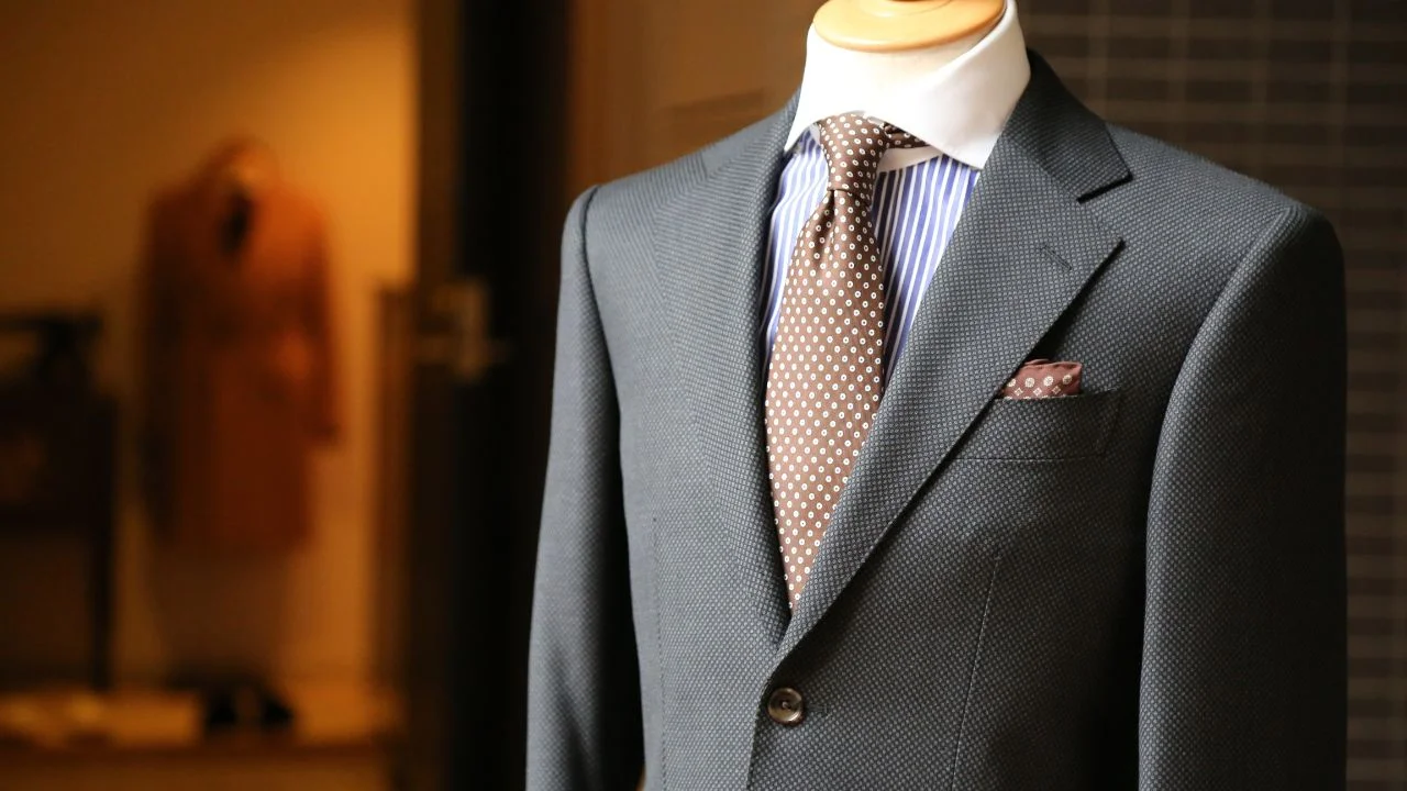 A dark gray suit jacket and pants on a mannequin. The suit has a white shirt, a blue and white striped tie, and a brown pocket square.