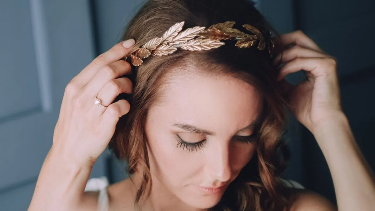 A woman adjusts a golden laurel wreath on her head, looking down with a soft expression. She is wearing a simple white top. Her fingers are delicately placed on her hair, and her face is soft and natural.