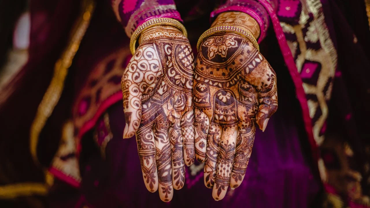 A close-up of a woman's hands adorned with intricate henna designs. The henna is dark brown and covers both of her palms and fingers.