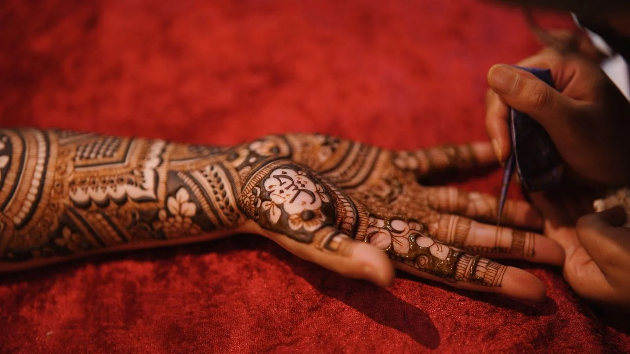 A close-up of a woman's hand with intricate Mehndi designs being applied by another person. The henna is dark brown and covers the woman's palm, fingers, and part of her wrist.