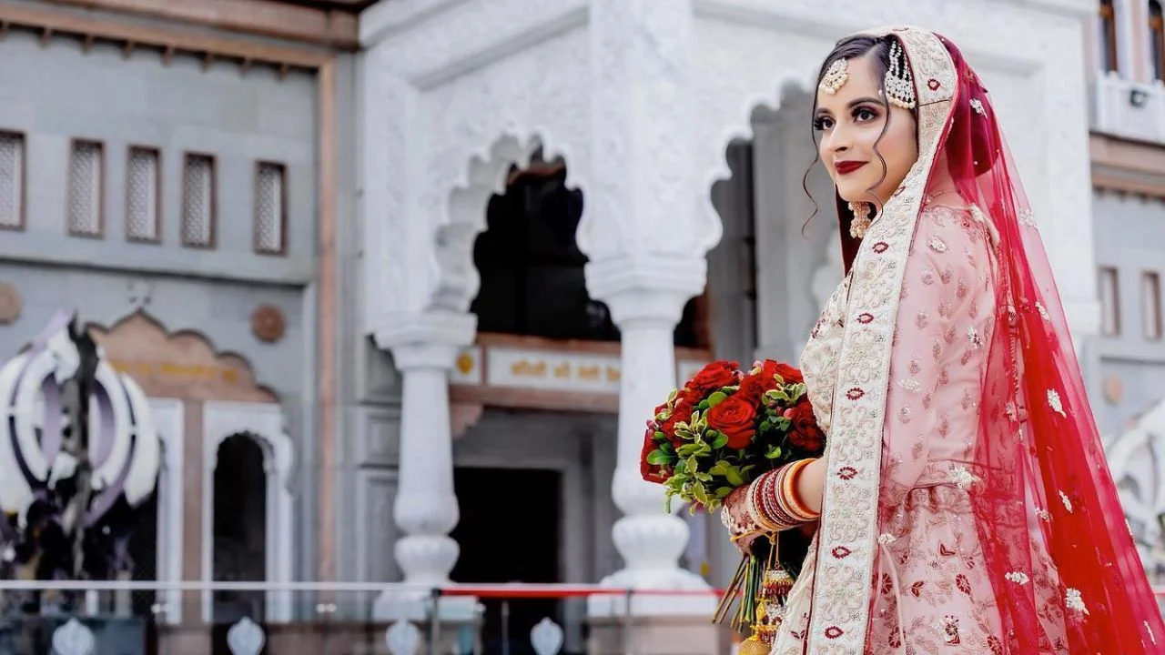 A bride in a red and white traditional Indian outfit with red dupatta stands in front of a white building, holding a bouquet of red roses.