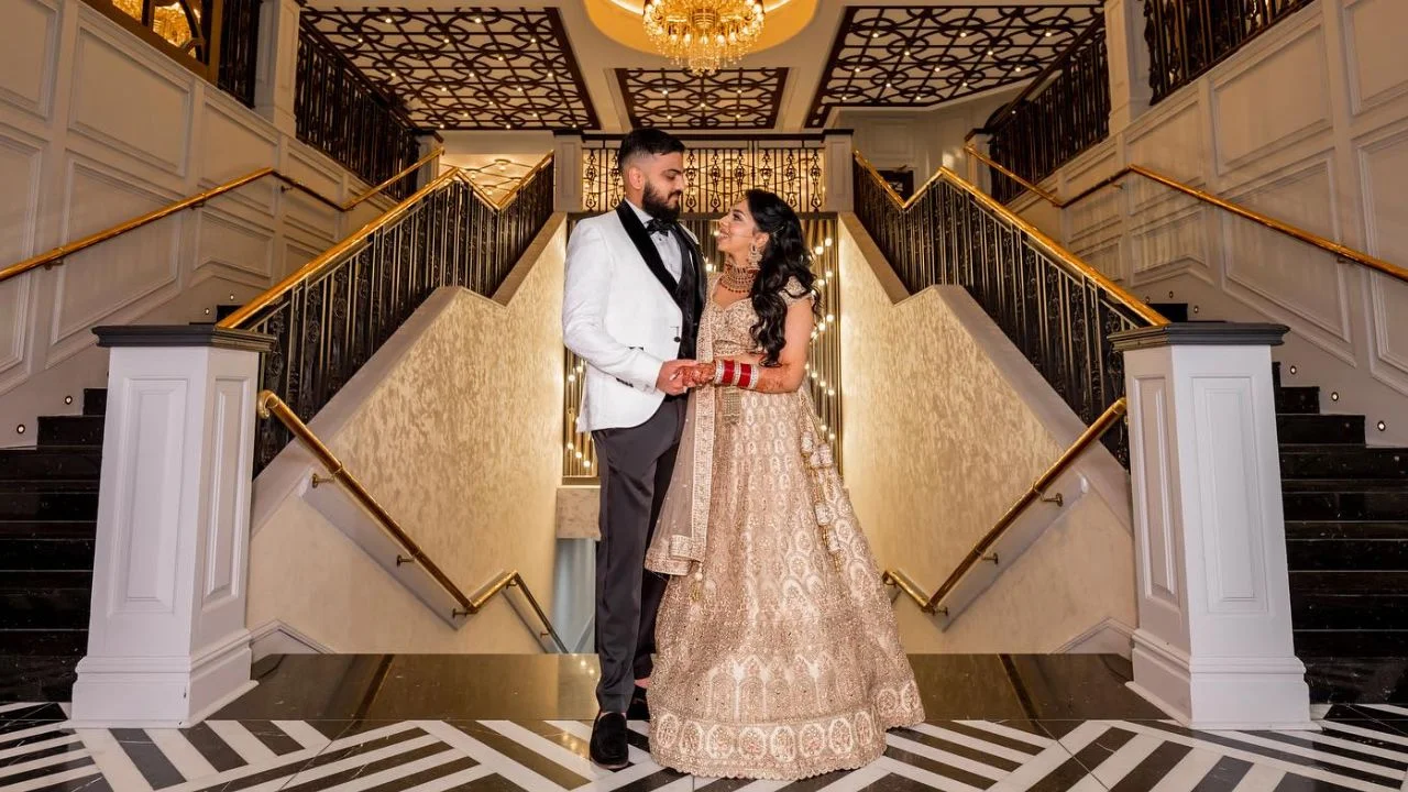 A newlywed couple poses on a grand staircase in an elegant venue. The groom is dressed in a white tuxedo jacket with black lapels, while the bride wears an ornate beige and gold lehenga adorned with intricate embroidery. They hold hands and gaze lovingly at each other, standing under a chandelier with beautifully patterned ceilings and stair railings, creating a luxurious and romantic atmosphere.